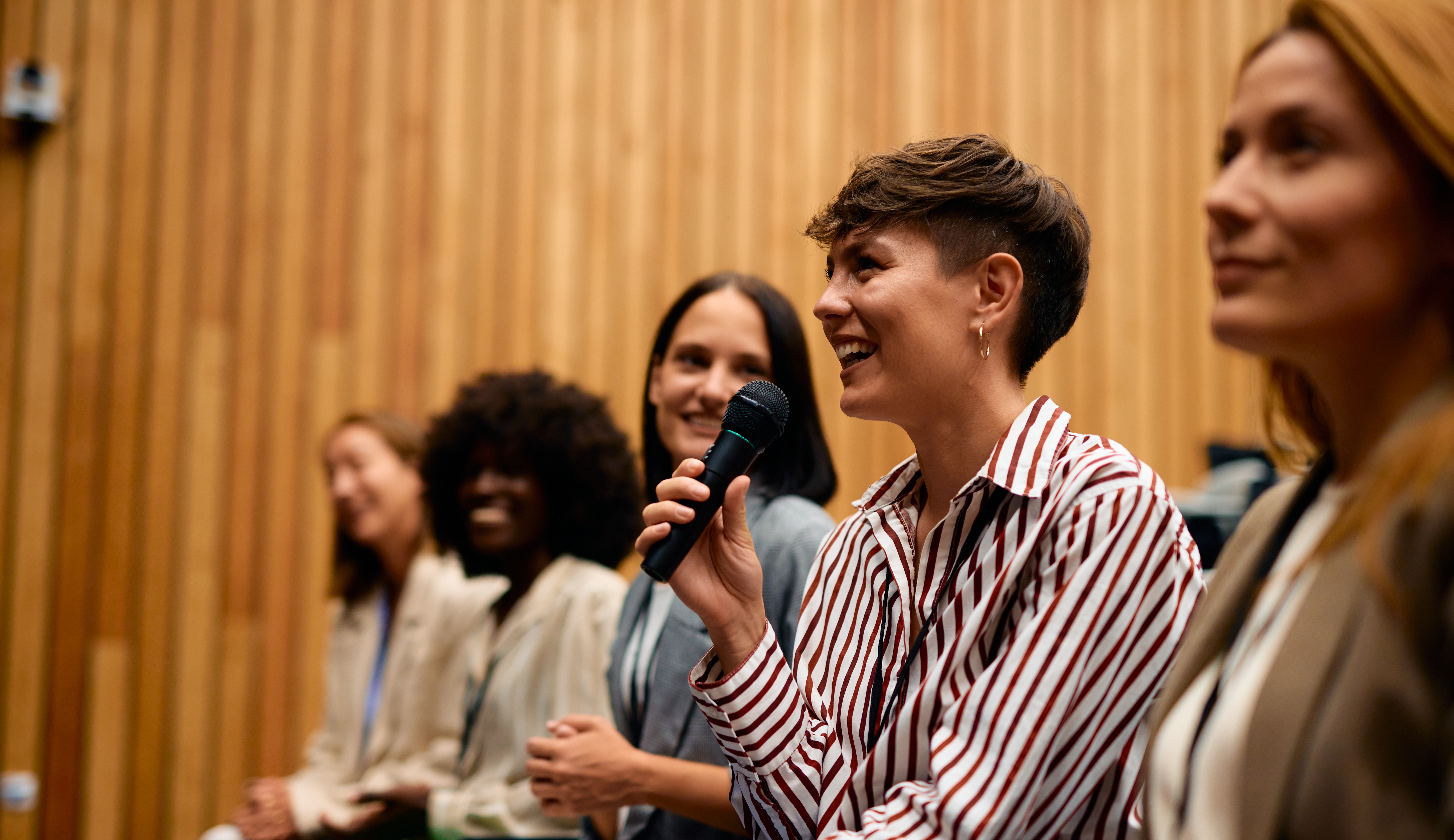 Photo of young business woman holding a microphone and smiling with other women sat next to her listening and smiling