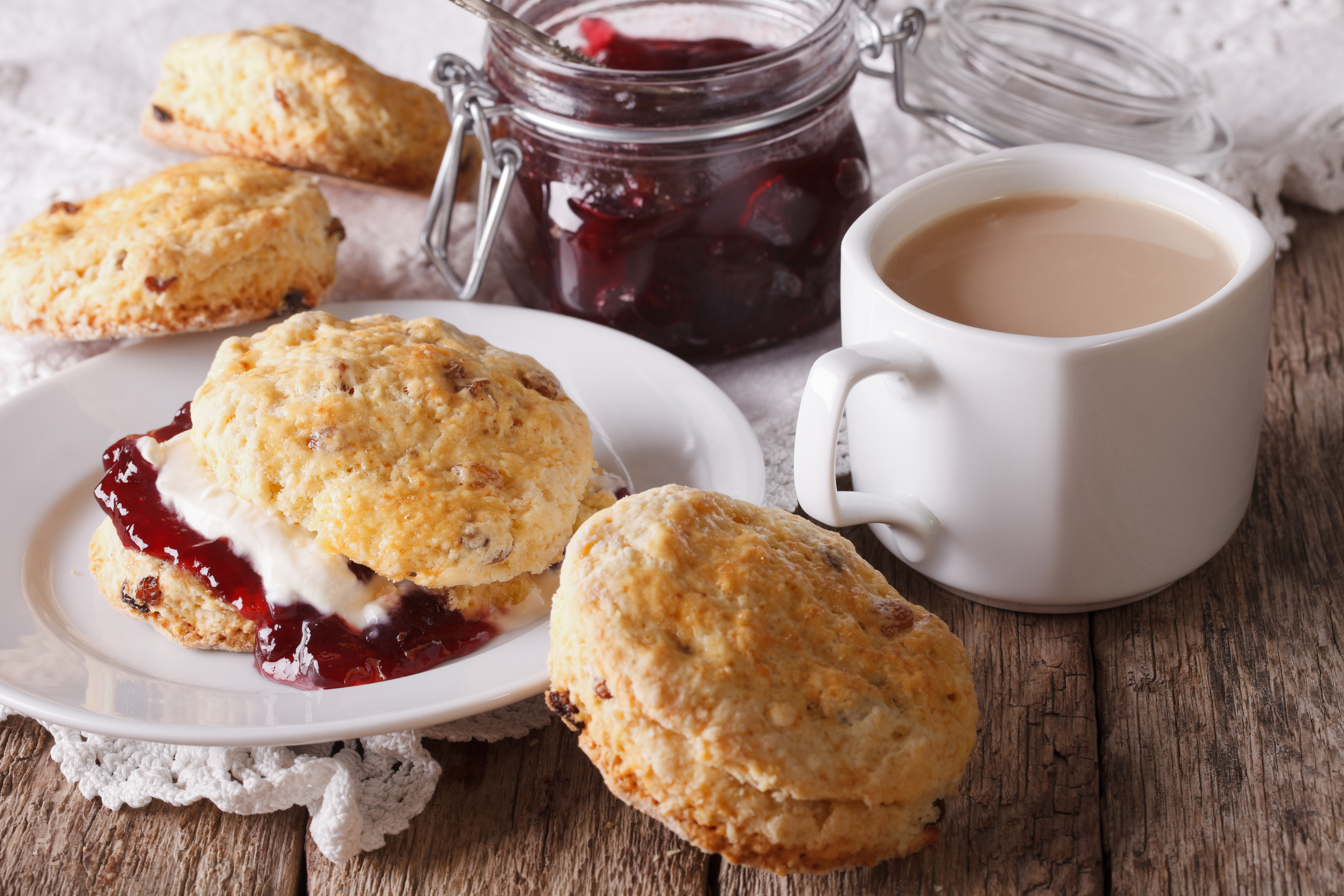 Scones filled with jam and cream sit on a plate alongside a cup of tea and a jar of jam.