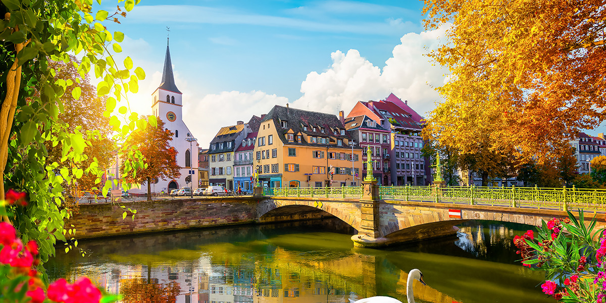 Strasbourg in the sunshine, with colour houses along a river, in the forefront a stone bridge