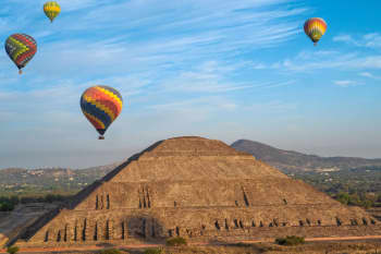 Three rainbow coloured hot air balloons soars across a blue sky under a huge stepped pyramid.