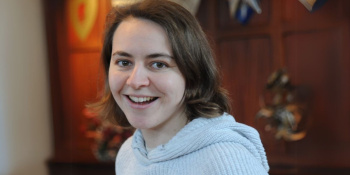 Headshot shows a female with chin length brown, wavy hair, wearing a grey woolly jumper.