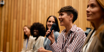 Photo of young business woman holding a microphone and smiling with other women sat next to her listening and smiling
