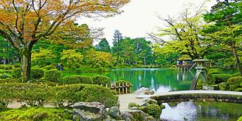 Lucious green Japanese garden with small concrete bridge over water with a Japanese house far in the background
