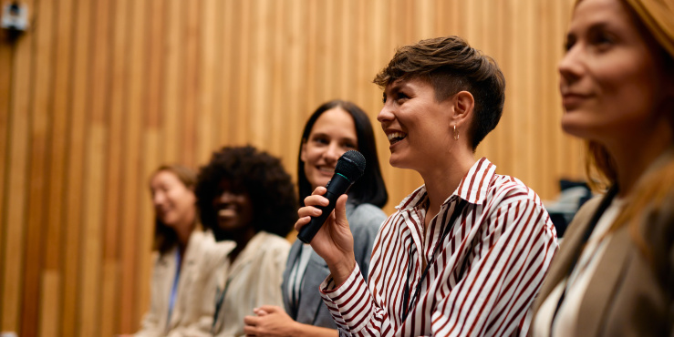 Photo of young business woman holding a microphone and smiling with other women sat next to her listening and smiling