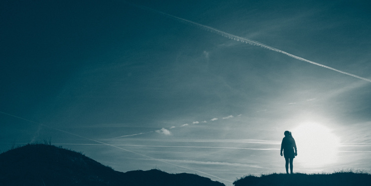 A woman stands at the top of a hill, looking out to the horizon on a sunny day.