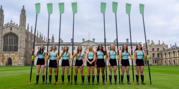 The Cambridge Women's Crew stand in a single line, holding their blades vertically towards the sky.