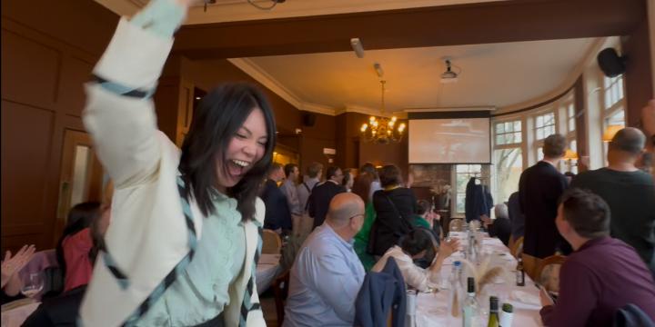 photograph of an alumna cheering in a room with multiple people watching the boat races