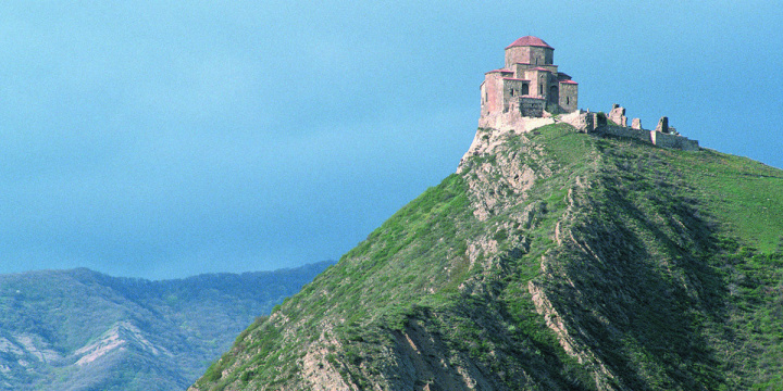 Sixth-century Georgian Orthodox monastery sitting on top of a green grass mountain with cloudy blue sky