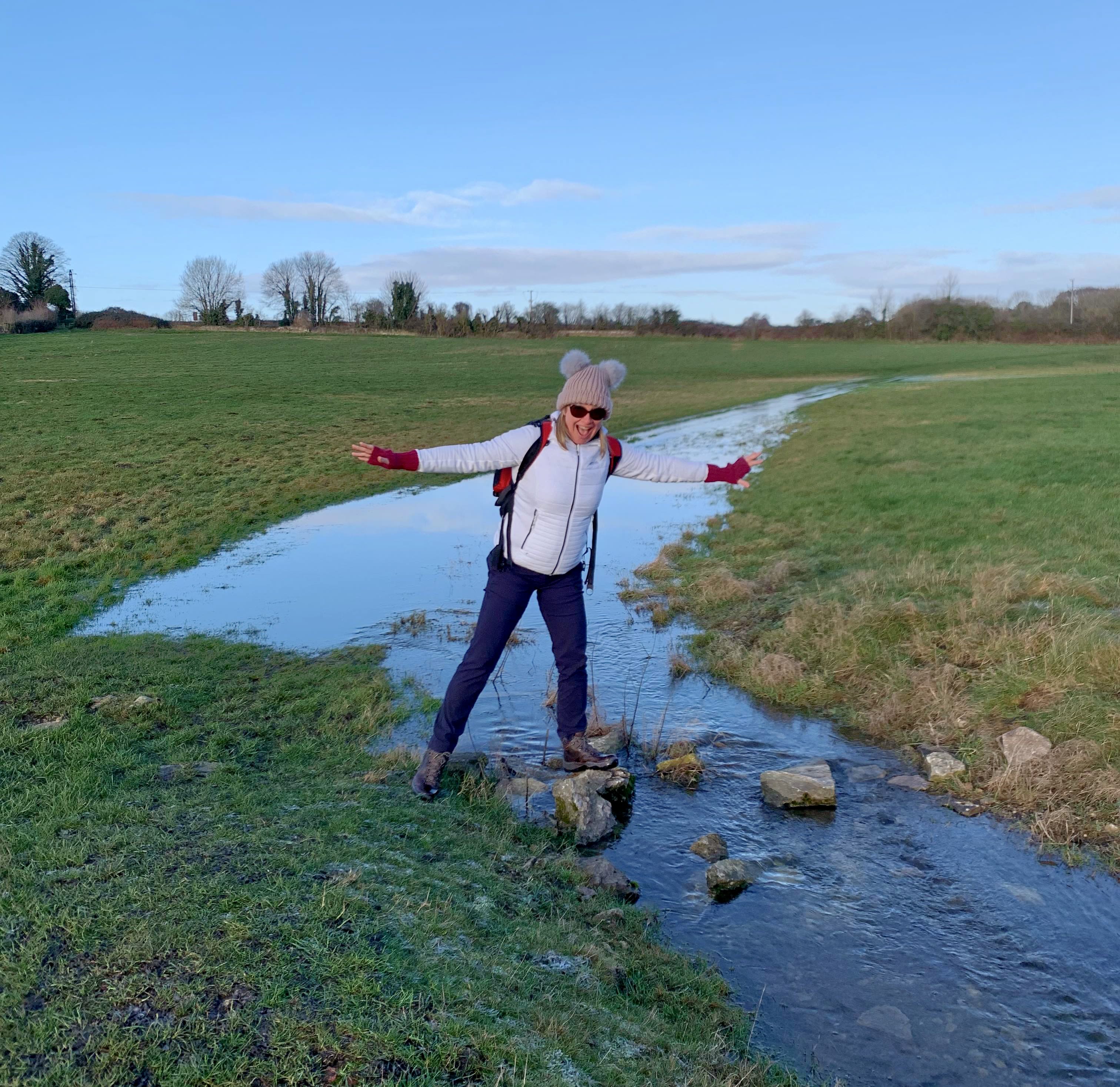 Claire stands in outdoor gear, balancing one one leg on a stepping stone in the middle of a stream.