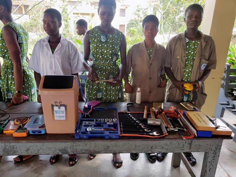 Tile shows 5 students dressed in beige and green school uniforms standing behind a desk on top of which electronic equipment is arranged.