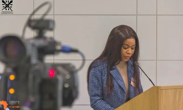 Alice stands at a podium in a blue suit jacket with white checks. In the foreground is a video camera recording her talk.