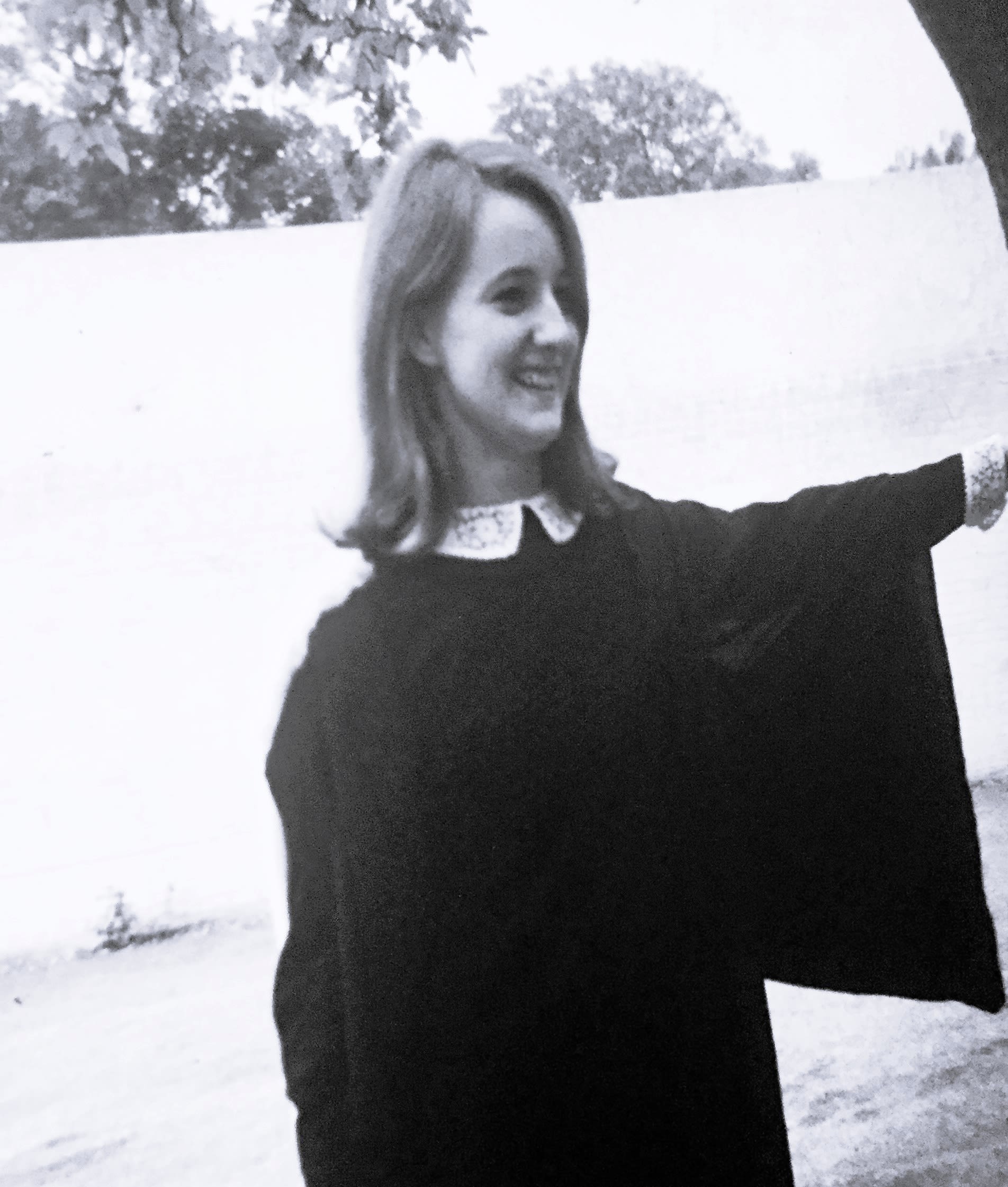 Katherine has shoulder length hair. She stands in a formal gown pointing and smiling.