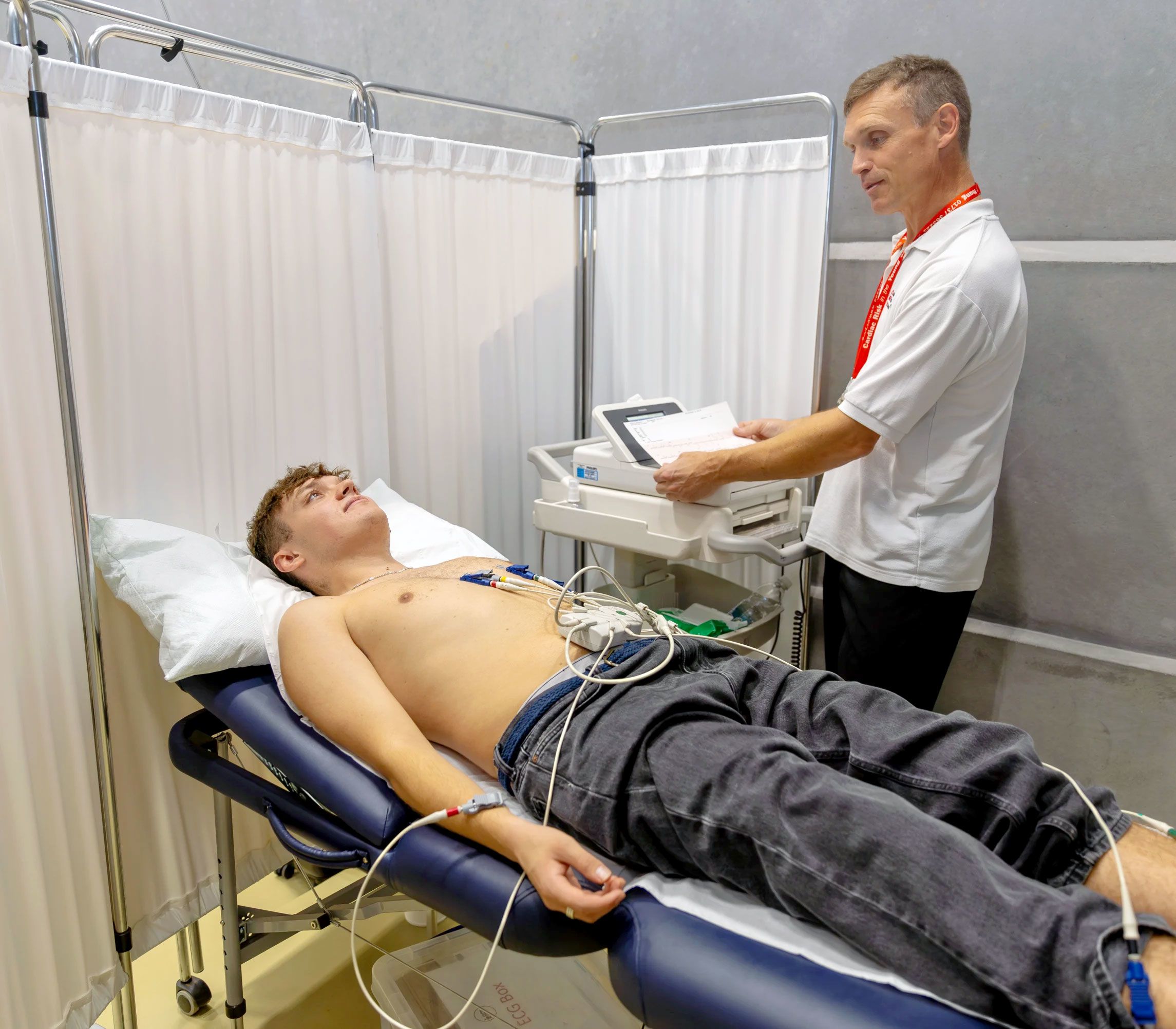 A young man lies shirtless on a bed whilst a male health care provided applied hear monitoring devices to his chest.