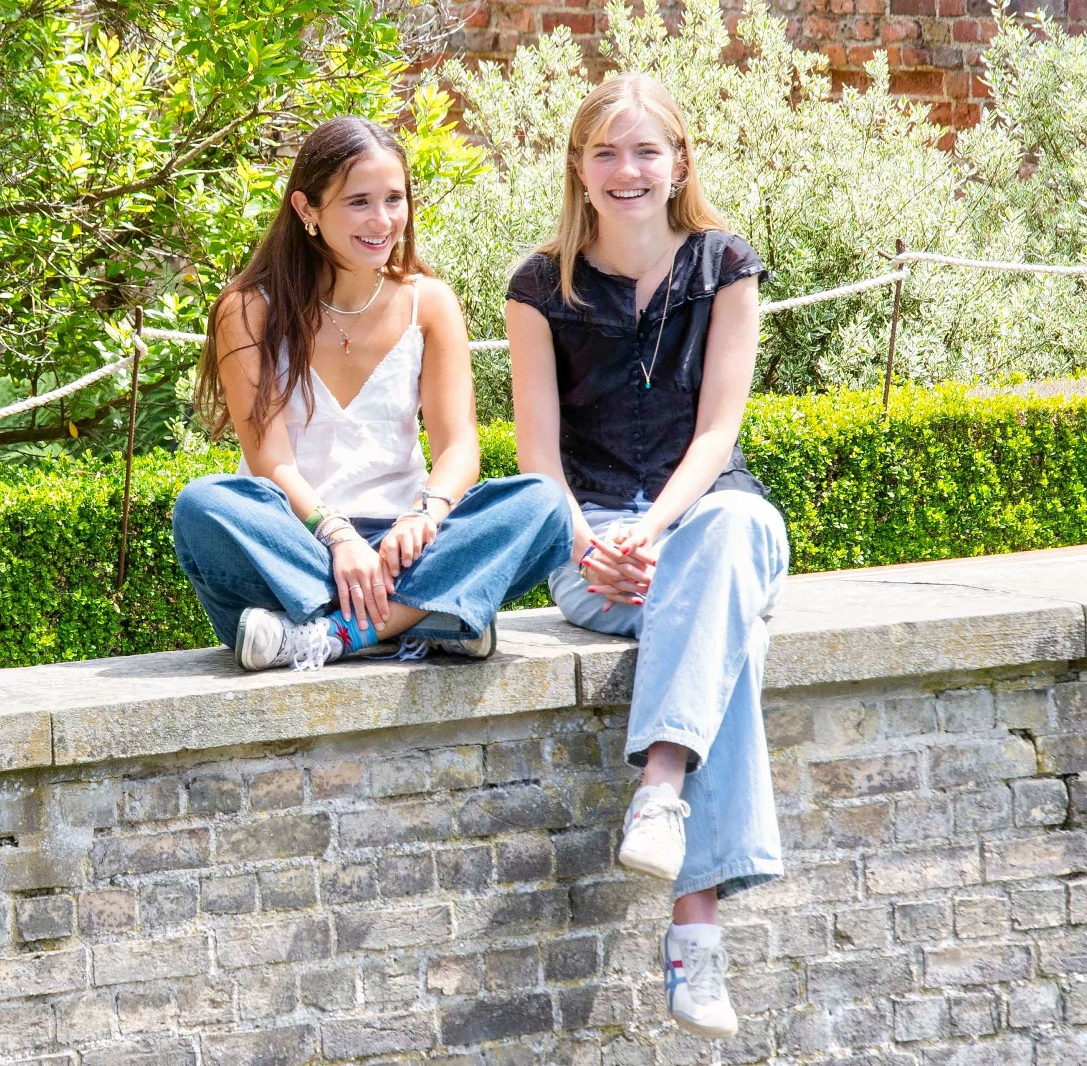 Two girls sit on a brick wall. One sits crossed-legged and the other has her legs dangling over the side. The both wear loose fitting jeans and light summer tops.