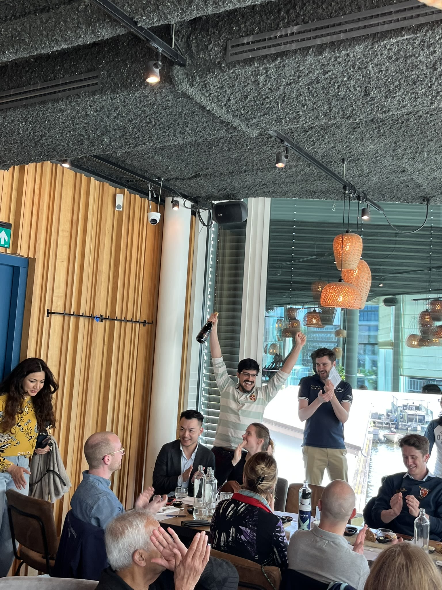 A man in a striped Light Blue top cheers with his hands in the air, holding a bottle of fizz in his right hand. His Oxford counterpart claps and smiles, standing to his left.