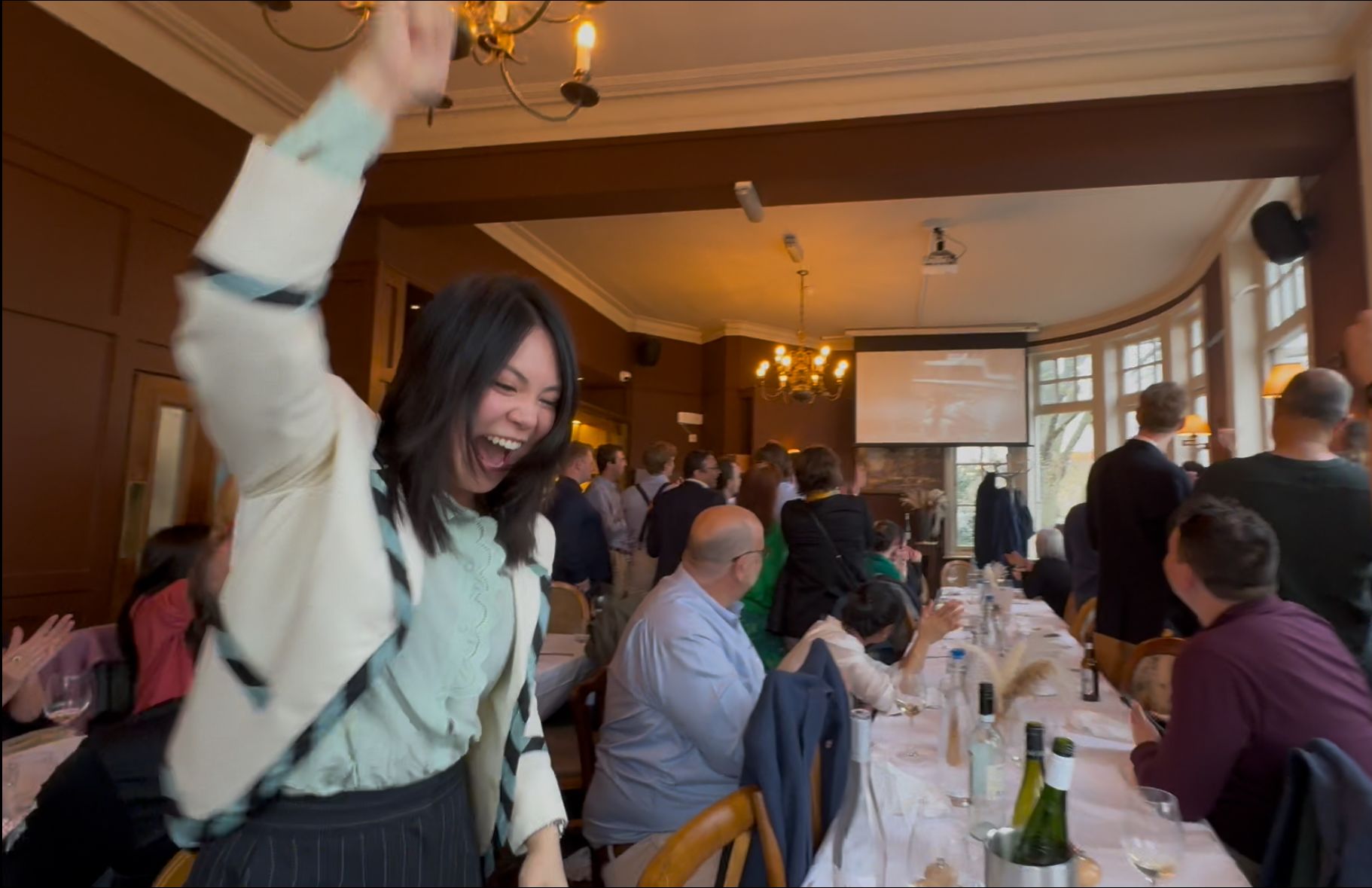 An alumna punches the air in joy. Behind her, people seated watch the Boat Race on a big screen. She's wearing a Light Blue jacket and top.