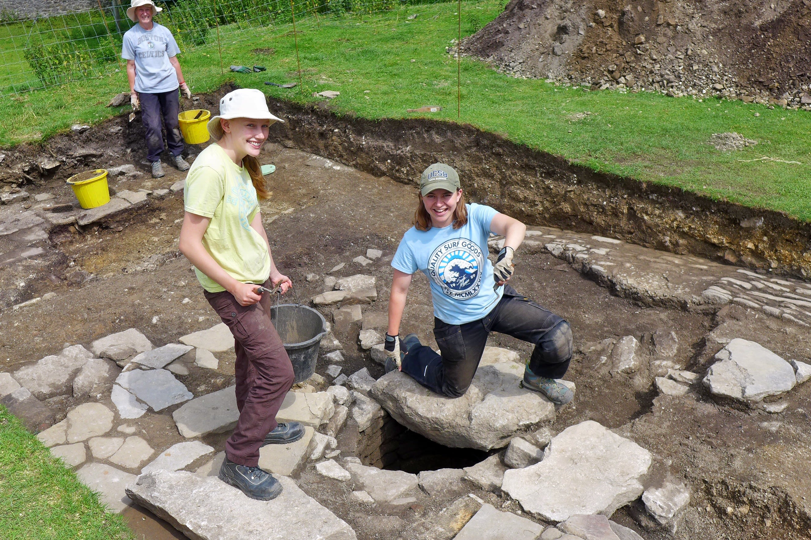 Morgause is one an archaeological site with two other archaeologists excavating a site. They each smile at the camera, holding trowels and buckets.