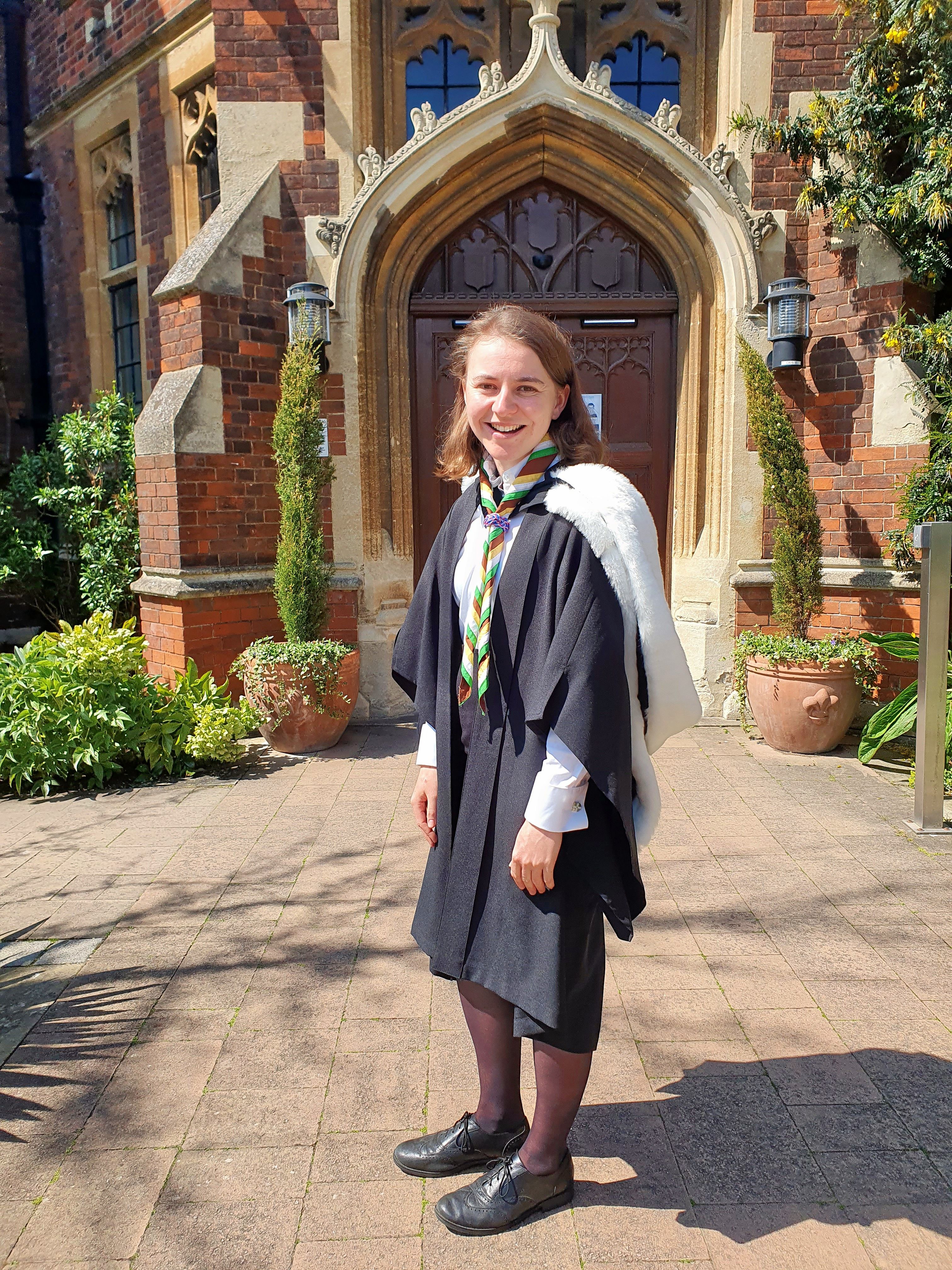 Morgause is in academic dress smiling in front of a historic building with ornate architecture. She has shoulder-length, light brown hair and wears a rainbow Scouting neckscarf.