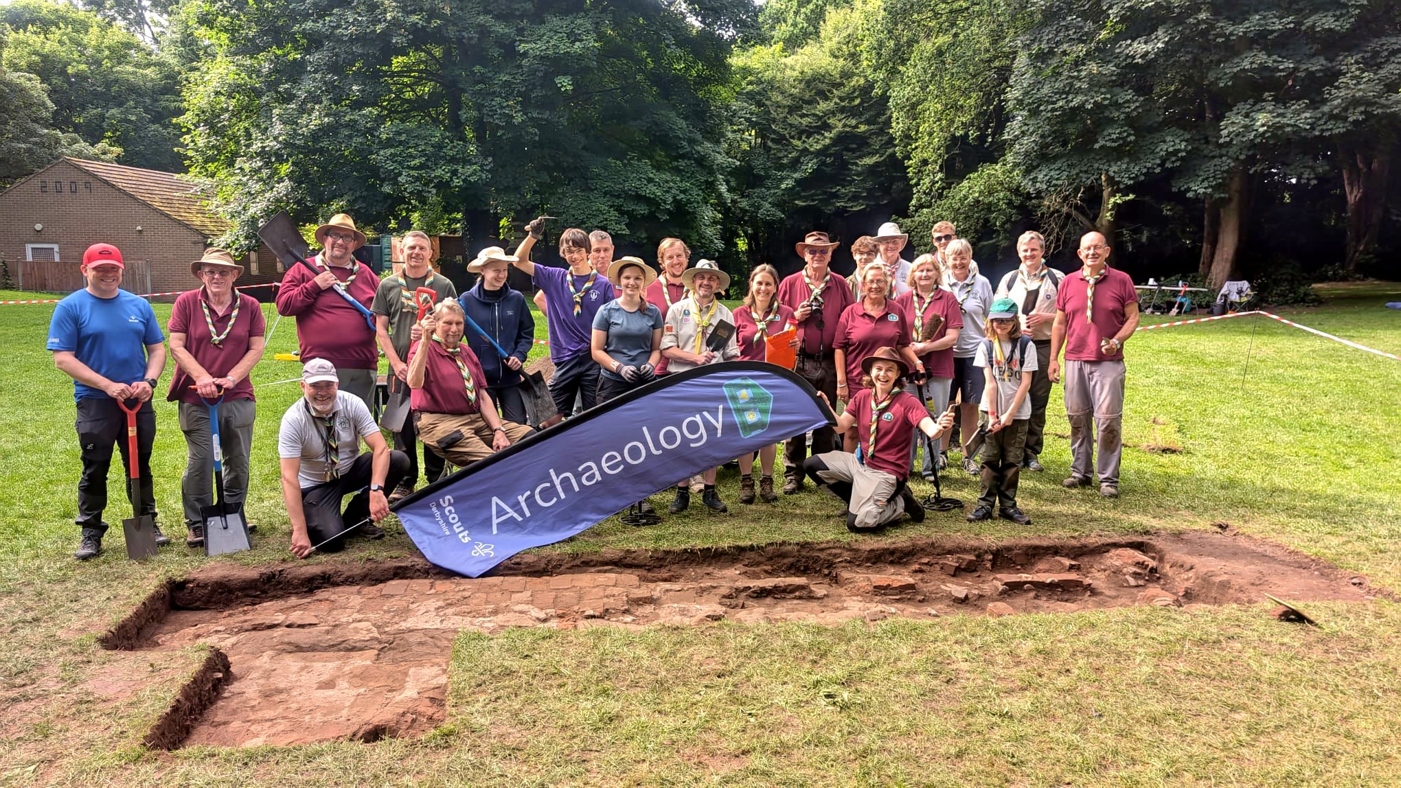 The Derbyshire Scout Archaeology Team are posing for a group photo, dressed in red shirts and green and yellow striped neck-scarfs. They are standing around an archaeological dig site, holding a banner that reads "Archaeology" and "Scouts".