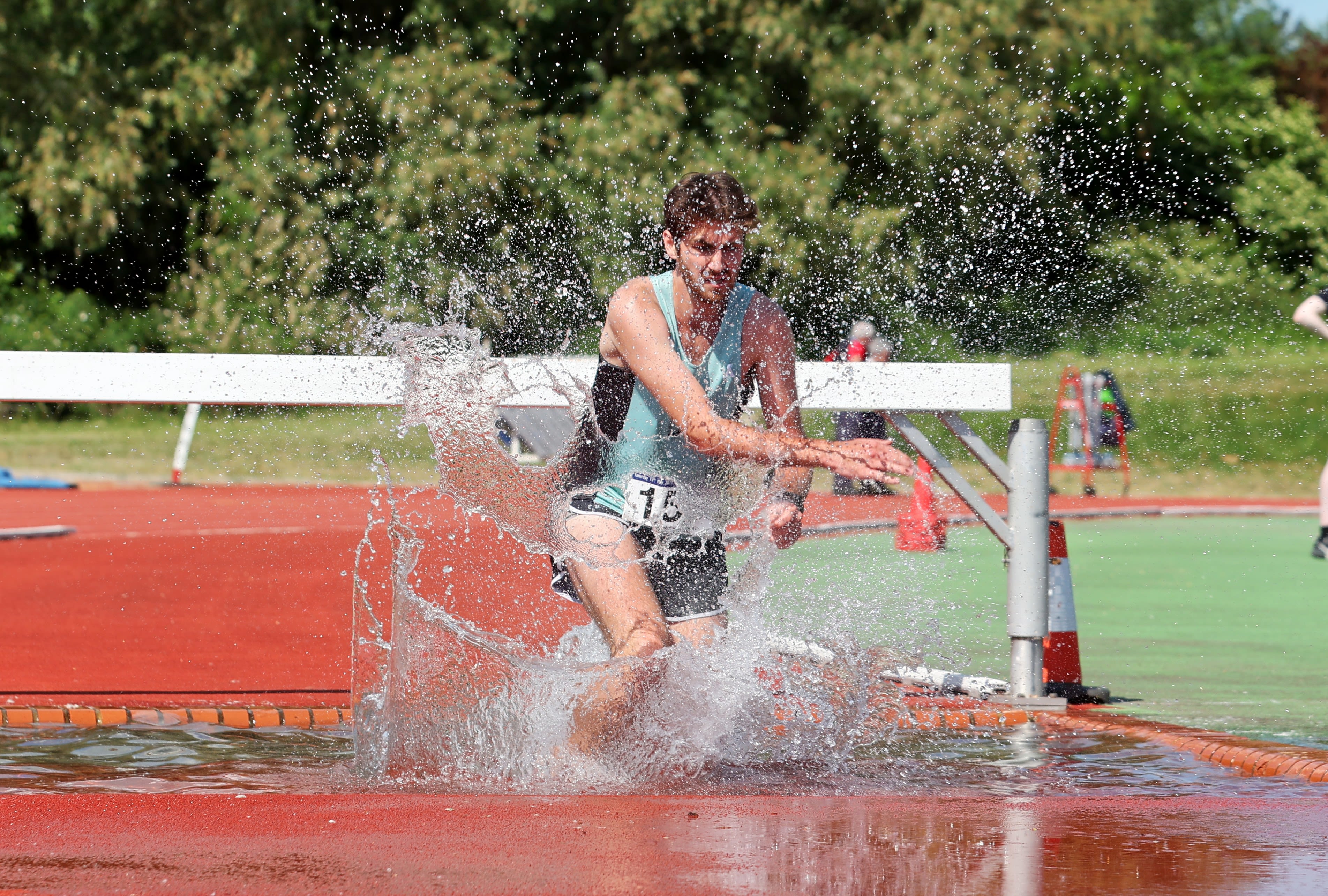 A male athlete lands in a pool of water as he clears a hurdle.