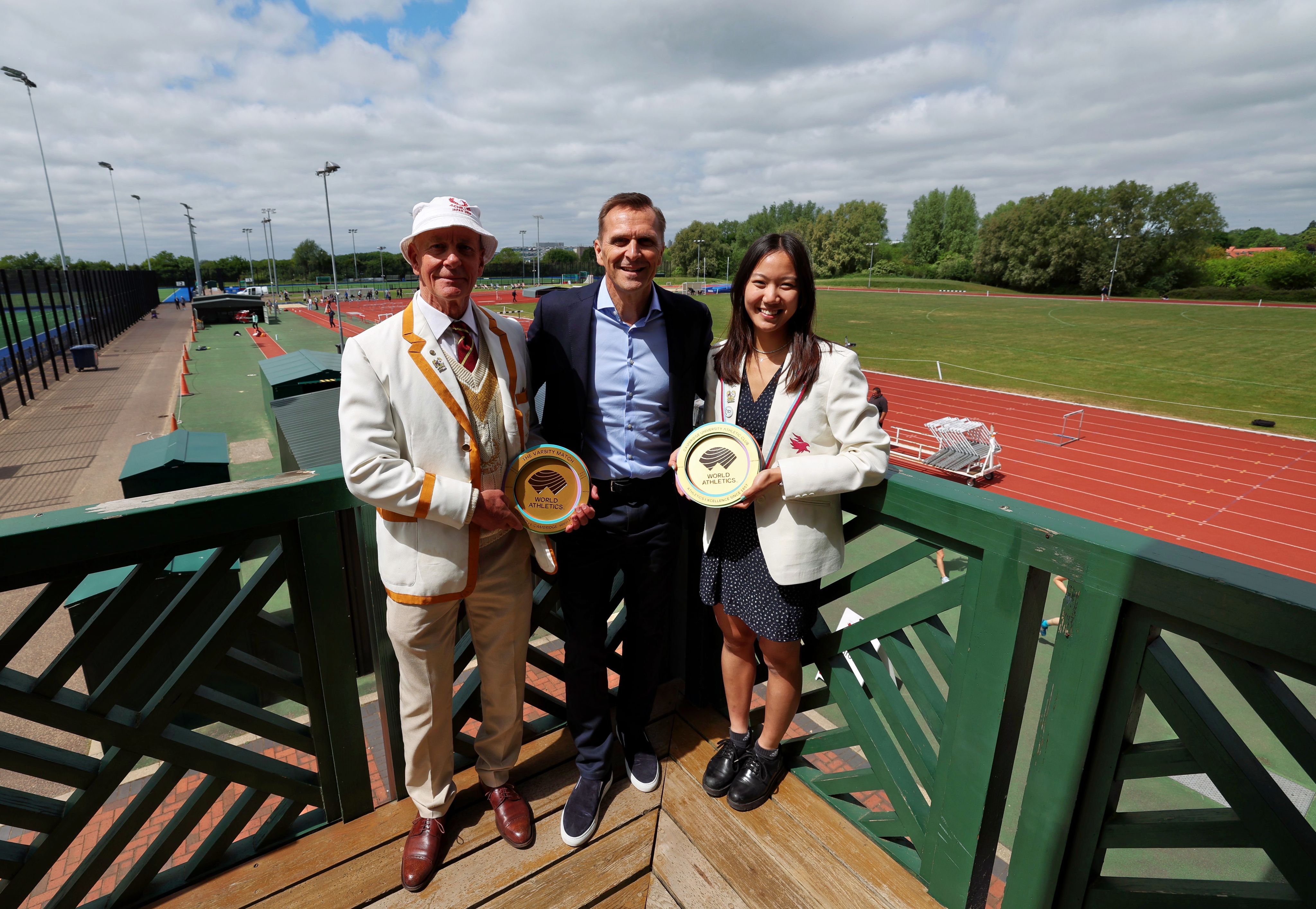 Three individual, two male and one female, stand on a balcony overlooking an athletics track holding golden disc-shaped plaques. The male on the left and the female on the right wear cream coloured Club blazers, while the central make wears a dark suit and light blue shirt.