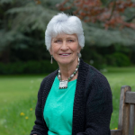 Profile picture of Sibella Laing, wearing a green t-shirt with a black cardy, sitting on a bench with green grass and trees