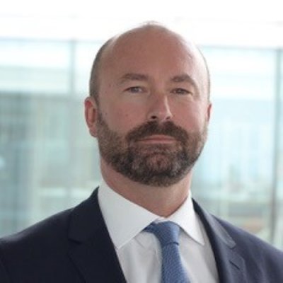 Portrait photograph of a beared man in a suit set in an office in front of glass panels