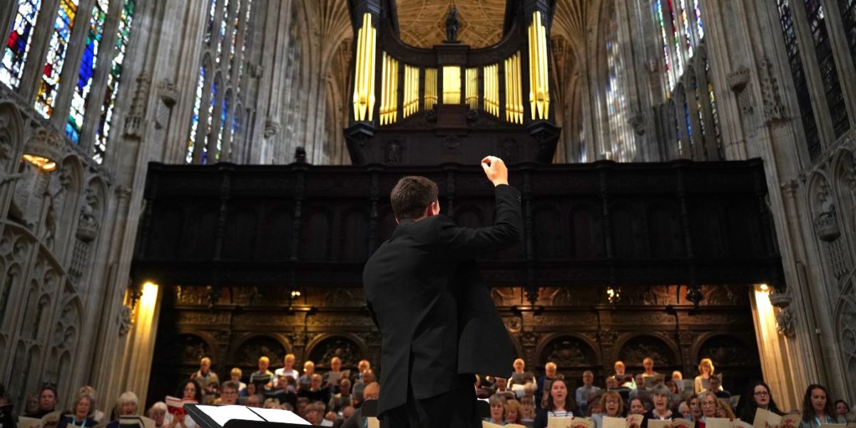A conductor stands with his back to us. In the background a choir sings in a chapel.