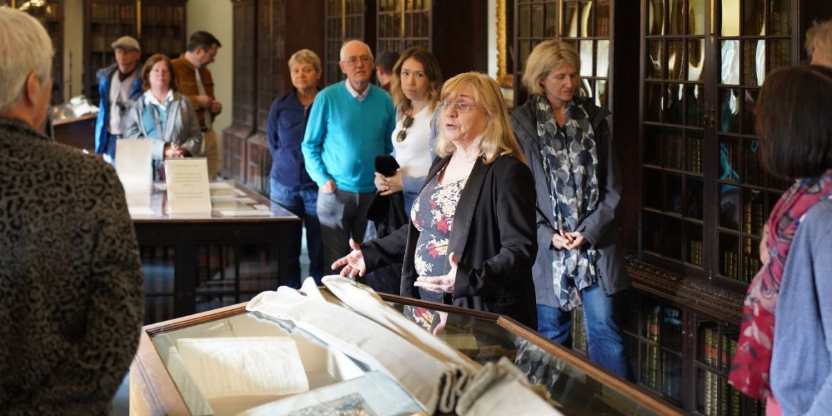 Visitors listen to a speaker in an ornate library with manuscripts laid out in central cases.