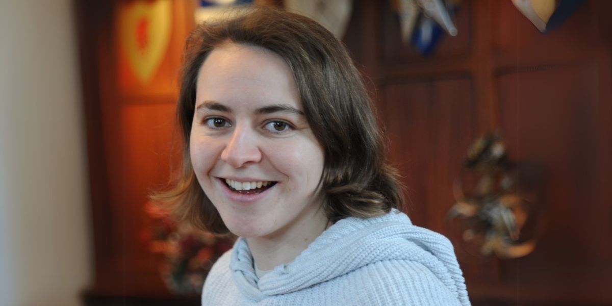 Headshot shows a female with chin length brown, wavy hair, wearing a grey woolly jumper.